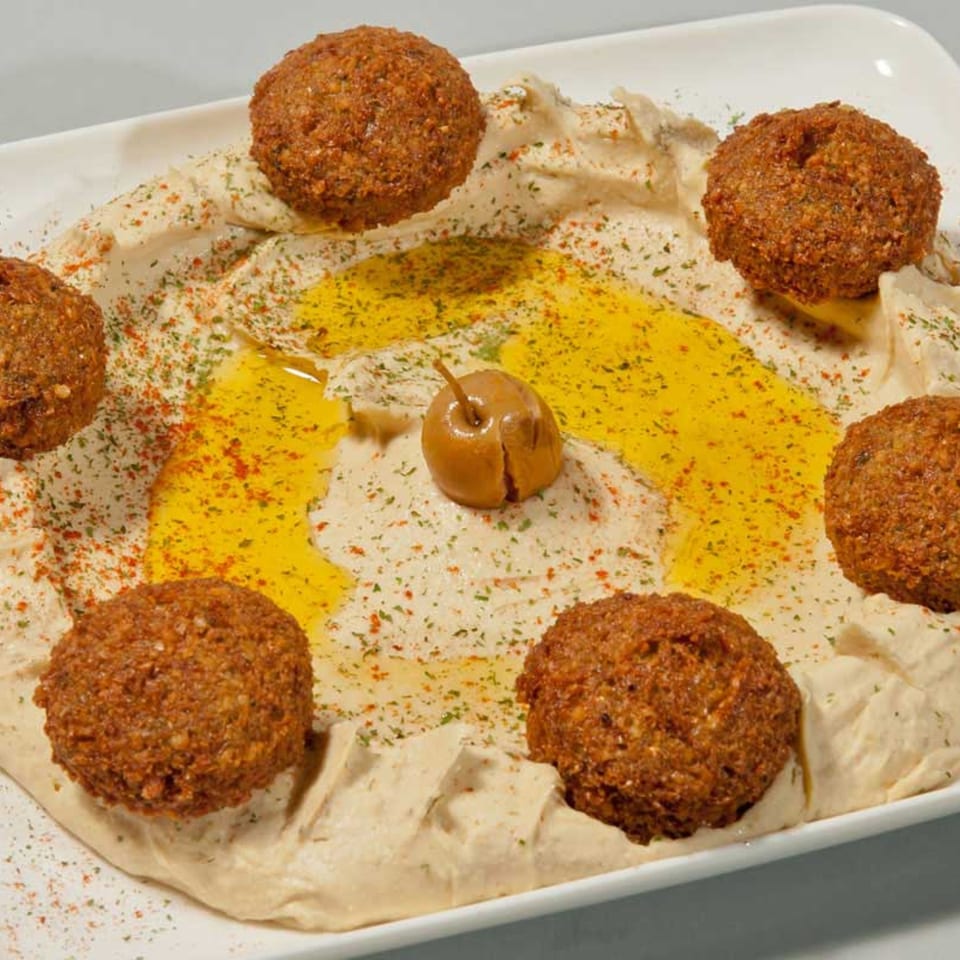 A white platter of hummus topped with falafel, olive oil, spices, and a single olive.