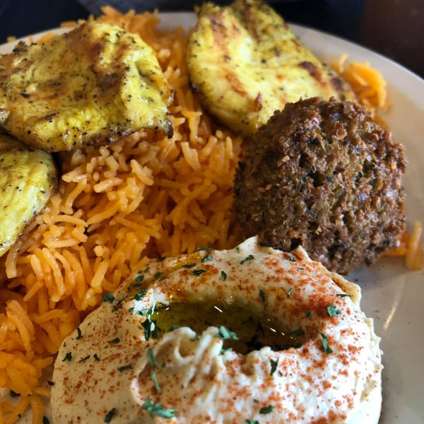 A Mediterranean platter with grilled chicken, orange rice, falafel, and hummus topped with paprika and parsley.