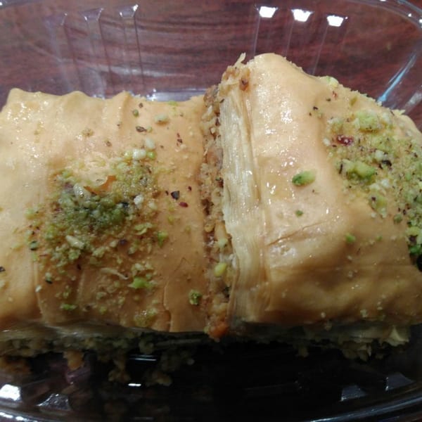 Two pieces of baklava, drizzled with syrup and topped with chopped pistachios, in a clear plastic container.